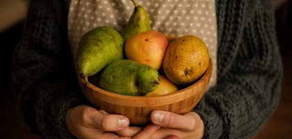 Close-up of hands holding a wooden bowl filled with pears and apples in warm indoor lighting.